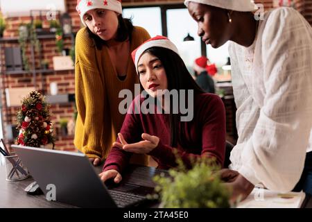 Diverse squadre femminili che indossano cappelli di babbo natale discutono di resoconti e brainstorming in un ufficio decorato di Natale. Colleghi che aiutano i dipendenti a svolgere attività durante le festività natalizie sul posto di lavoro Foto Stock