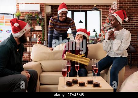 Una donna asiatica impiegata nel cappello di babbo natale seduta sul divano dell'ufficio e con in mano una confezione regalo di Natale da parte dei colleghi. Diversi colleghi che giocano babbo natale segreto e condividono regali alla festa di Capodanno Foto Stock