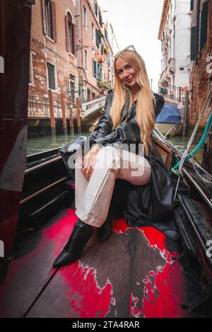 Elegante donna con giacca e pantaloni in pelle nera che si gode un giro in gondola attraverso i pittoreschi canali di Venezia. Foto Stock