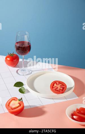 Pomodori freschi, piatti in ceramica bianca e un bicchiere di vino rosso sono posti sul tavolo. Sfondo blu e rosa. La vitamina C nei pomodori aiuta a stimolare il colon Foto Stock