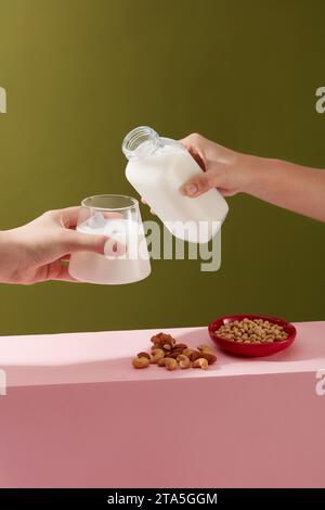 Due mani con una tazza di vetro e una bottiglia di latte. Piatto rosso contenente soia posto accanto a una manciata di mandorle e noci di anacardi. I dadi contengono unsat Foto Stock