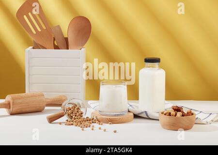 Vista frontale di una tazza per il latte posizionata su un podio di legno, esposta con una ciotola di noci, semi di soia, utensili da cucina e una bottiglia di latte. Flacone etichetta vuoto per p Foto Stock