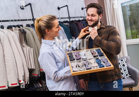 Venditrice che indossa una cravatta su un cliente, entrambe sorridenti, porta abiti dietro in un negozio di abbigliamento maschile Foto Stock