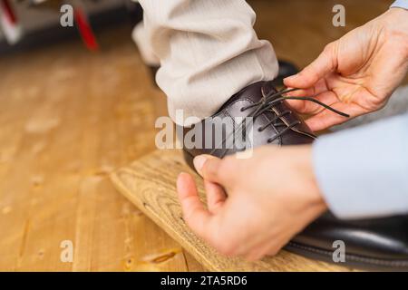 Primo piano di mani che legano i lacci di una nuova scarpa marrone in pelle su un tabellone di legno in un negozio di matrimoni Foto Stock