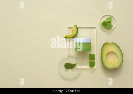 Sullo sfondo beige, barattolo cosmetico e crema su podi di vetro decorati con fette di avocado fresche. Il concetto di cosmetici naturali biologici Foto Stock