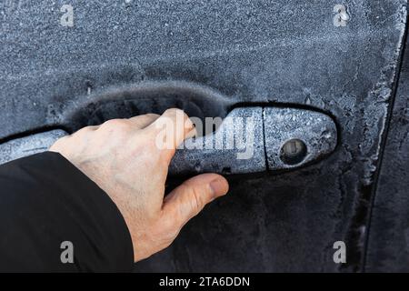 Mano che cerca di aprire la porta chiusa a chiave di un'auto nera coperta di gelo nella stagione invernale fredda Foto Stock