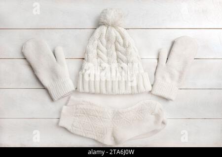 Un cappello in maglia bianca con pompon, mittensa di lana e calzini giacciono su uno sfondo di legno bianco. Dettagli dell'abbigliamento invernale, piatto, vista dall'alto Foto Stock