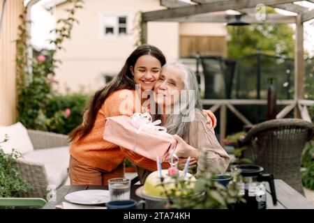 Felice ragazza che abbraccia e dà un regalo di compleanno alla nonna nel patio Foto Stock