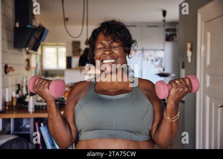 Donna matura che stringe i denti mentre si esercita con i manubri a casa Foto Stock