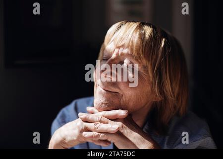Donna anziana bionda sorridente con le mani sul mento che si rilassa alla luce del sole a casa Foto Stock