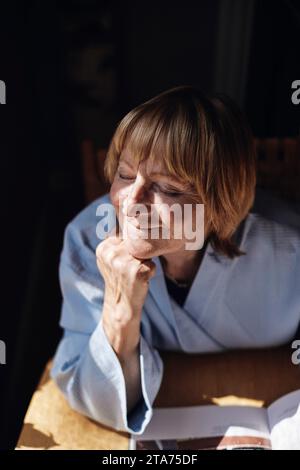 Donna anziana bionda con la mano sul mento che si rilassa alla luce del sole a casa Foto Stock