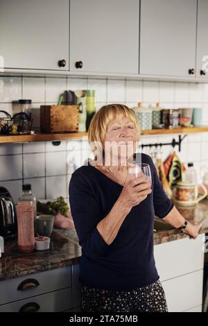 Donna anziana bionda con gli occhi chiusi che beve un bicchiere in cucina a casa Foto Stock