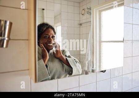 Donna matura felice che tocca il volto mentre si guarda nello specchio del bagno a casa Foto Stock