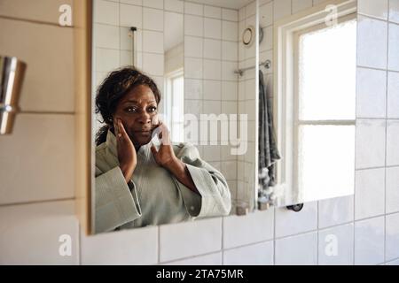 Donna matura che tocca il volto mentre guarda nello specchio in bagno a casa Foto Stock