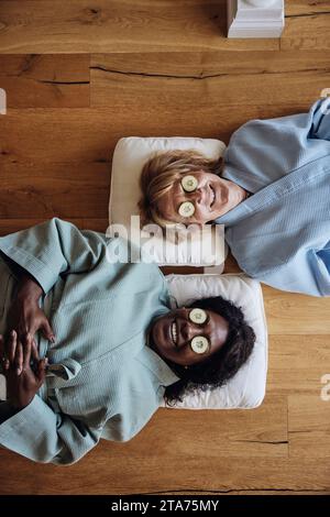 Direttamente sopra la vista delle amiche sorridenti con fette di cetriolo sugli occhi che si rilassano sul pavimento a casa Foto Stock