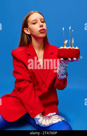 Ritratto di una giovane donna vestita abiti insoliti, stravaganti e luminosi, indossa collant blu, giacca rossa con torta di compleanno guardando la macchina fotografica. Foto Stock