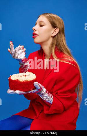 Ritratto di una ragazza travestita, stravagante, insolita e vivida seduta e che mangia con le mani una deliziosa torta di compleanno. Foto Stock