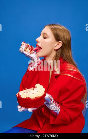 Ritratto di una ragazza felicissima, stravagante, insolita e vivida seduta e leccata di torta dolce e gustosa su sfondo blu. Foto Stock