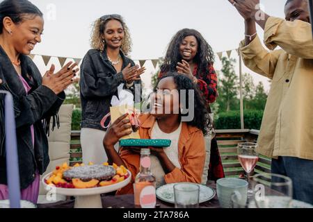 Amici allegri che applaudono una donna sorridente che regala regali durante la festa di compleanno nel cortile posteriore Foto Stock