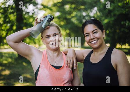 Ritratto di una donna sorridente con un'amica esausta che tiene una bottiglia d'acqua al parco Foto Stock