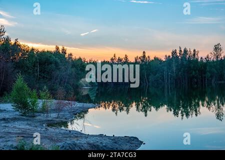 Questa immagine cattura la bellezza serena di un lago nella foresta al crepuscolo. Le acque calme creano uno specchio perfetto, riflettendo gli alberi fitti e la sfumatura dei colori del cielo al tramonto. La luce soffusa sul bordo della giornata crea un'atmosfera tranquilla e la delicata transizione dalla terra all'acqua sul bordo del lago invita a una tranquilla contemplazione. Questo è un momento di simmetria naturale, dove il confine tra terra e cielo sembra dissolversi nella quiete della sera. Twilight Reflections al Forest Lake. Foto di alta qualità Foto Stock