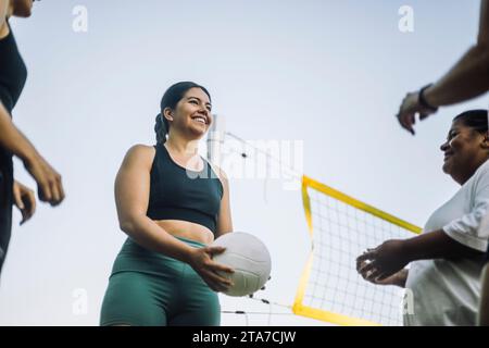 Vista dall'angolo basso di una donna sorridente che gioca a pallavolo con gli amici contro il cielo Foto Stock