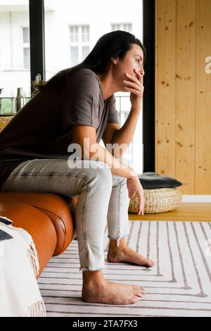 Donna preoccupata con la bocca che copre le mani seduta sul divano a casa Foto Stock