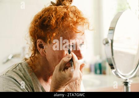 Donna anziana che guarda allo specchio e applica l'eyeliner a casa Foto Stock