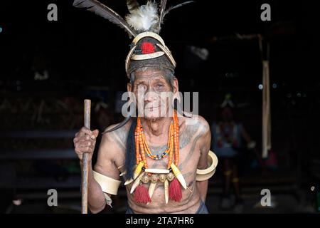 Anziano della tribù indigena Konyak in abiti tradizionali con cappello tradizionale sulla testa seduto in una casa di legno fatta di bambù, Nagaland, India Foto Stock