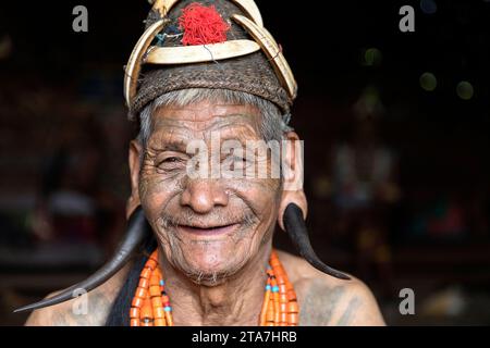 Guerriero anziano della tribù Konyak in abiti tradizionali con faccia tatuata in una casa di legno fatta di bambù, Nagaland, India Foto Stock