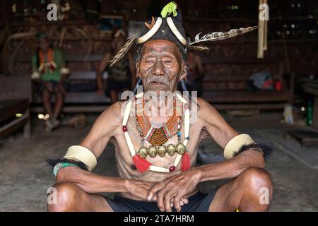 Anziano della tribù indigena Konyak in abiti tradizionali con cappello tradizionale sulla testa seduto in una casa di legno fatta di bambù, Nagaland, India Foto Stock