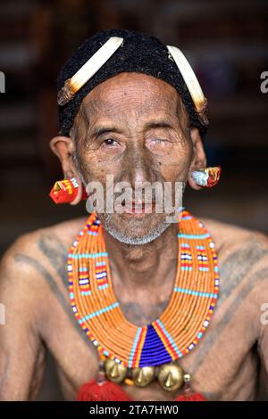 Anziano della tribù indigena Konyak in abiti tradizionali con cappello tradizionale sulla testa seduto in una casa di legno fatta di bambù, Nagaland, India Foto Stock