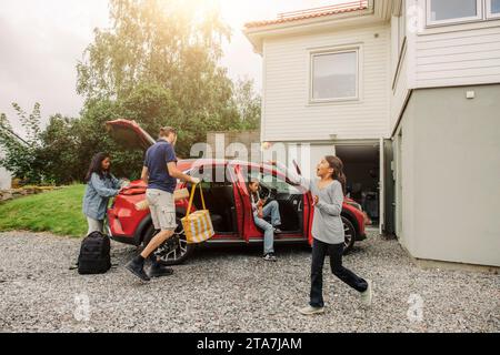 Ragazza che gioca con la palla vicino a tutta la famiglia caricando oggetti in auto elettrica a casa Foto Stock