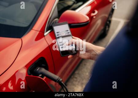 Vista ad alto angolo dell'uomo che utilizza l'app sullo smartphone vicino all'auto elettrica Foto Stock