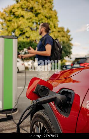 Auto di ricarica con cavo elettrico con uomo in piedi sullo sfondo alla stazione Foto Stock