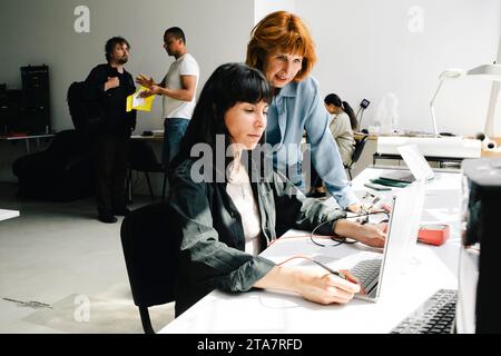 Donna anziana vicino a un tecnico donna che misura la tensione del computer portatile utilizzando un multimetro presso un'officina di riparazione Foto Stock