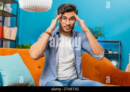 Malato Indiano indù che sfrega le tempie per curare il problema del mal di testa, soffre di tensione e emicrania, stress, dolore, pressione alta a casa. Un uomo arabo al coperto in camera sul divano Foto Stock