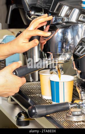 Le mani del barista controllano la macchina del caffè, l'espresso versa il caffè in due tazze di carta. Immagine verticale. Foto Stock