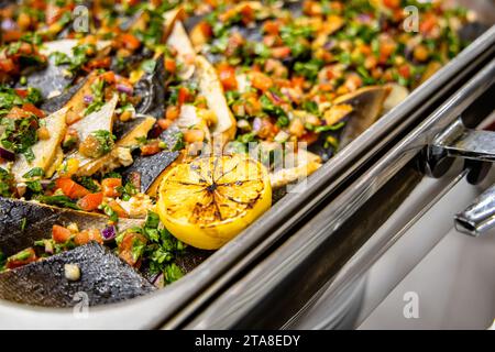 Pesce alla griglia con limone e verdure su un vassoio di metallo in un ristorante. ristorazione a buffet al coperto in un ristorante di lusso con salmone frutta colorata Foto Stock