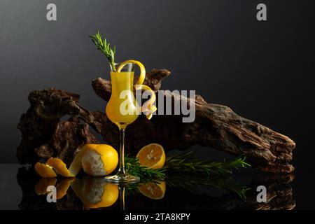 Liquore tradizionale italiano Limoncello su sfondo nero riflettente. Bicchiere di liquore al limone con ramoscello di rosmarino su sfondo di vecchio serpente. Foto Stock