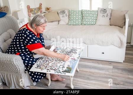 Una donna anziana che raccoglie puzzle mentre è seduta su una sedia nella sua stanza luminosa a casa. Foto Stock