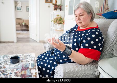 Donna anziana che misura la pressione sanguigna e il polso utilizzando il tonometro elettronico medico mentre è seduta su una poltrona in una stanza luminosa. Una donna anziana sovrappeso e problemi di ipertensione. Foto Stock