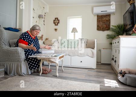 Una donna anziana che raccoglie puzzle mentre è seduta su una sedia nella sua stanza luminosa a casa. Piccolo cane che dorme in un cestino morbido. Foto Stock