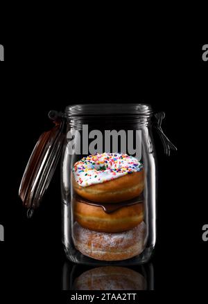Tre ciambelle in un contenitore in vetro o in un recipiente di inscatolamento isolato su nero con riflesso, con coperchio aperto. Foto Stock
