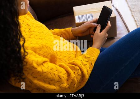 Sezione centrale della donna birazziale in maglione giallo con pacchetto che utilizza lo smartphone a casa Foto Stock