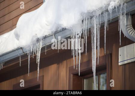 Ghiaccioli cristallini e luccicanti che si sono formati a causa delle condizioni climatiche gelide, appesi al bordo di una residenza in legno marrone Foto Stock