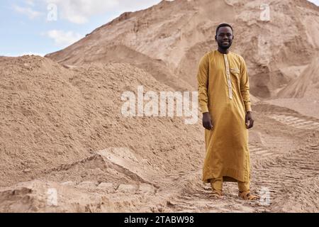 Ritratto grandangolare di un giovane uomo nero che indossa abiti tradizionali e sorride davanti alla macchina fotografica in piedi nel deserto, nello spazio per le copie Foto Stock