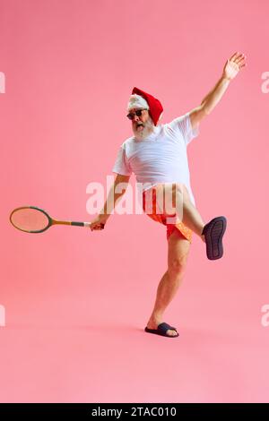 Ritratto a grandezza naturale di Babbo Natale vestito per le vacanze estive trascorrono le vacanze invernali giocando lo sport preferito, tennis su sfondo da studio color rosa pastello. Foto Stock