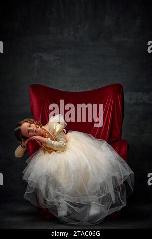 Graziosa bambina seduta in trono rosso vestita come una persona medievale in un abito vecchio stile e sognando su uno sfondo vintage scuro. Foto Stock