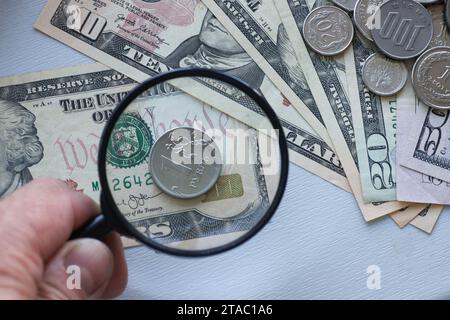 Lente d'ingrandimento, denaro e sanzioni finanziarie. Una mano regge una lente d'ingrandimento sopra un rublo russo di metallo sullo sfondo delle banconote da dollaro. Foto Stock
